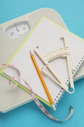 Body fat caliper, measuring tape, scale and notebook on light blue background, closeup