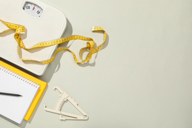 Body fat caliper, measuring tape, scale and notebook on light grey background, flat lay. Space for text