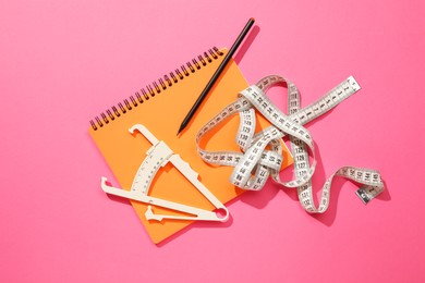 Body fat caliper, measuring tape, notebook and pencil on pink background, flat lay