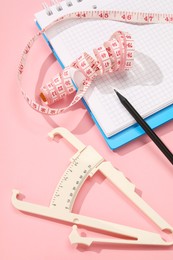 Photo of Body fat caliper, measuring tape, notebook and pencil on pink background