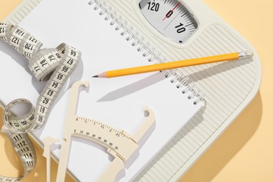 Photo of Body fat caliper, measuring tape, scale, pencil and notebook on beige background, top view