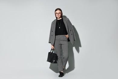 Beautiful woman in stylish suit and glasses with bag on gray background