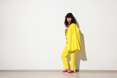 Beautiful young woman in stylish yellow suit near light wall, space for text