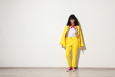 Beautiful young woman in stylish yellow suit near light wall, space for text