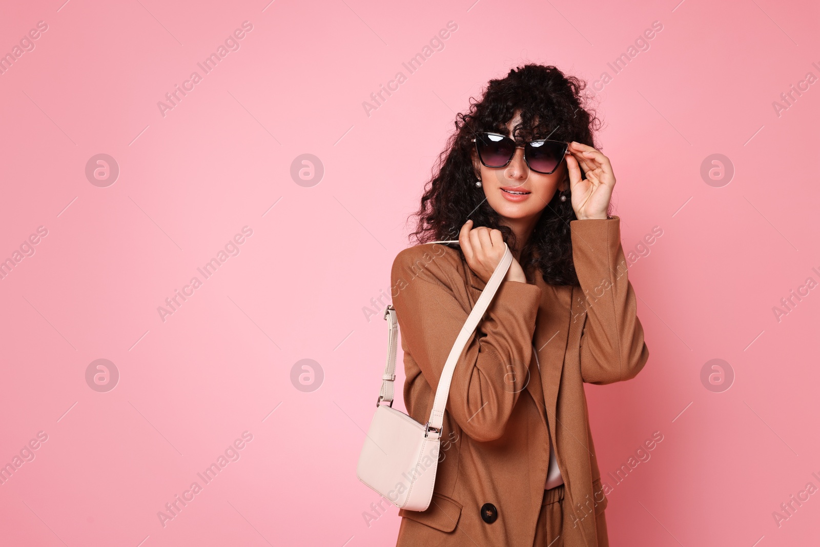 Photo of Beautiful young woman in stylish suit and sunglasses with bag on pink background, space for text