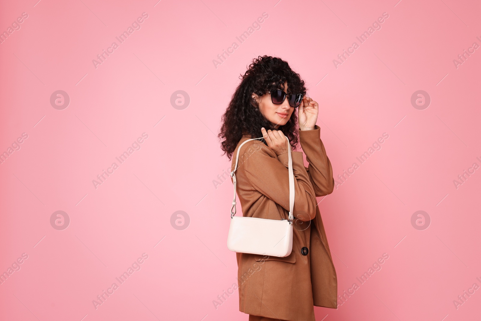 Photo of Beautiful young woman in stylish suit and sunglasses with bag on pink background