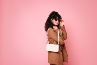 Beautiful young woman in stylish suit and sunglasses with bag on pink background