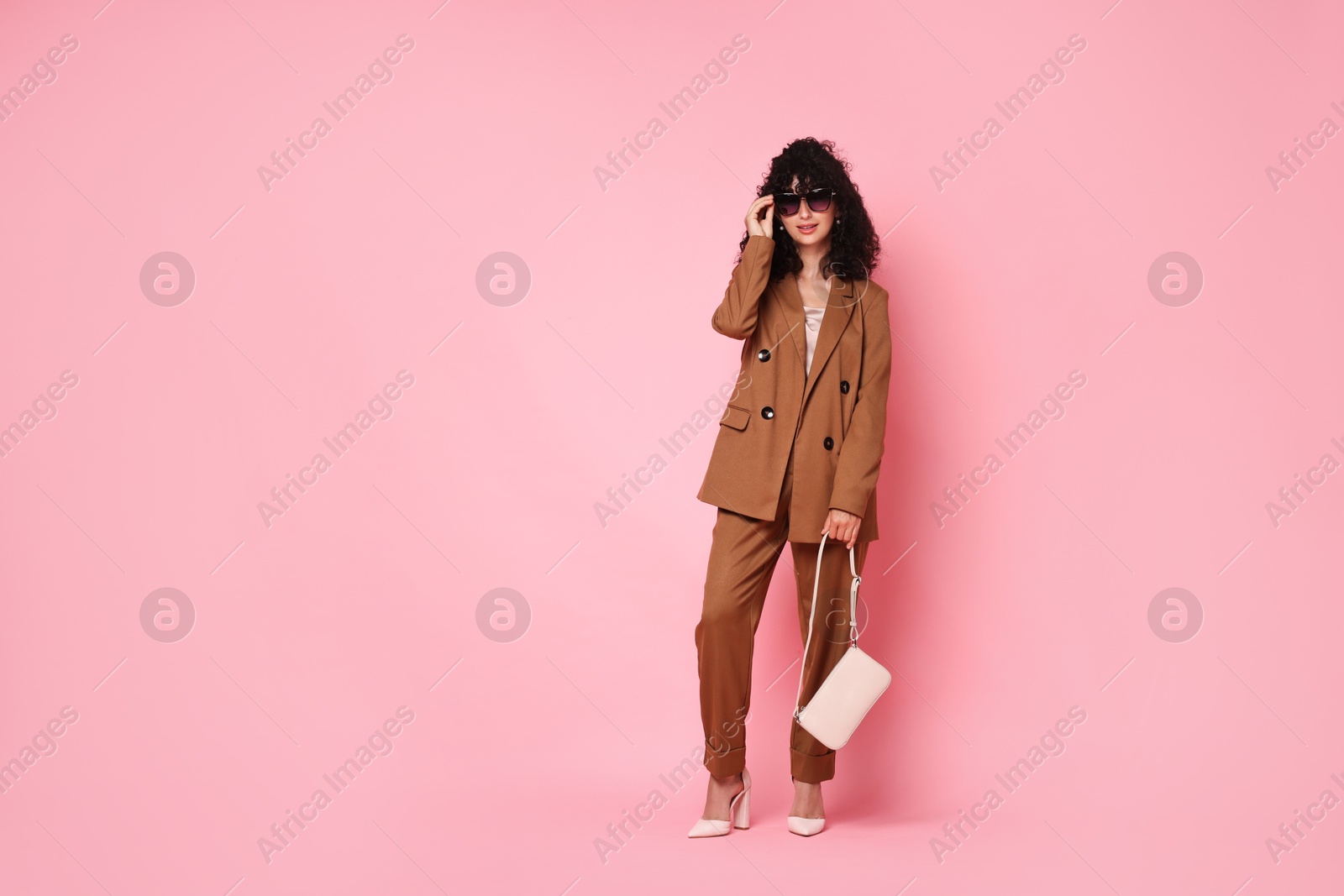 Photo of Beautiful young woman in stylish suit and sunglasses with bag on pink background