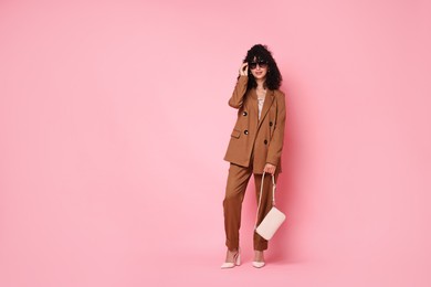 Beautiful young woman in stylish suit and sunglasses with bag on pink background