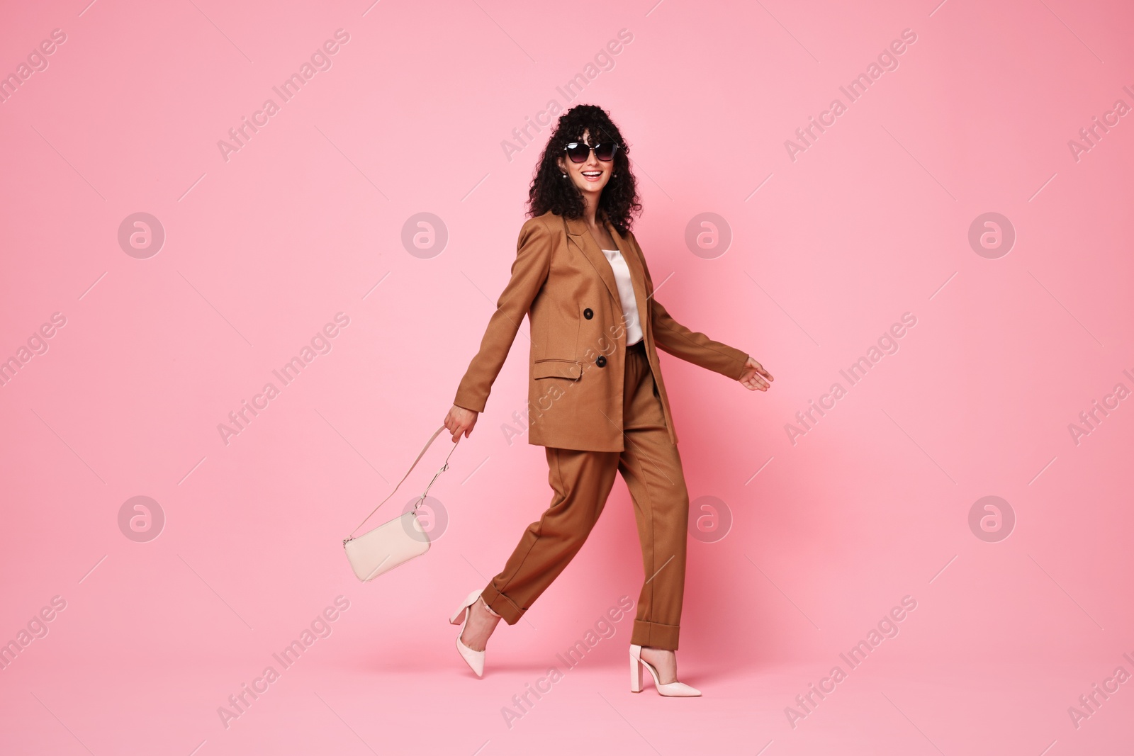 Photo of Beautiful young woman in stylish suit and sunglasses with bag walking on pink background