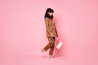 Photo of Beautiful young woman in stylish suit and sunglasses with bag walking on pink background