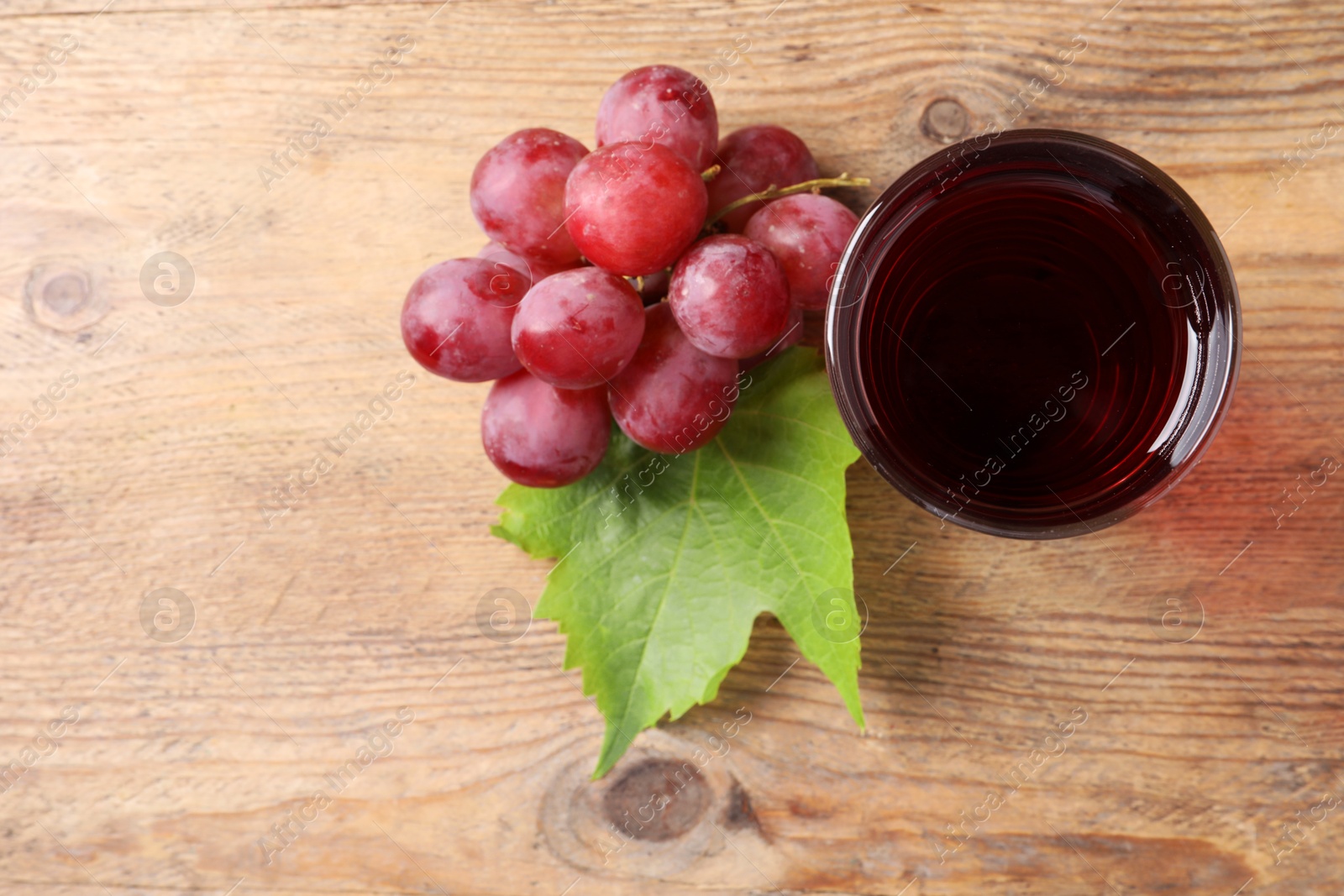 Photo of Tasty juice in glass, fresh grapes and leaf on wooden table, top view. Space for text