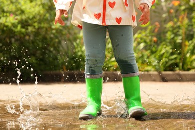 Photo of Little girl wearing green rubber boots standing in puddle outdoors, closeup