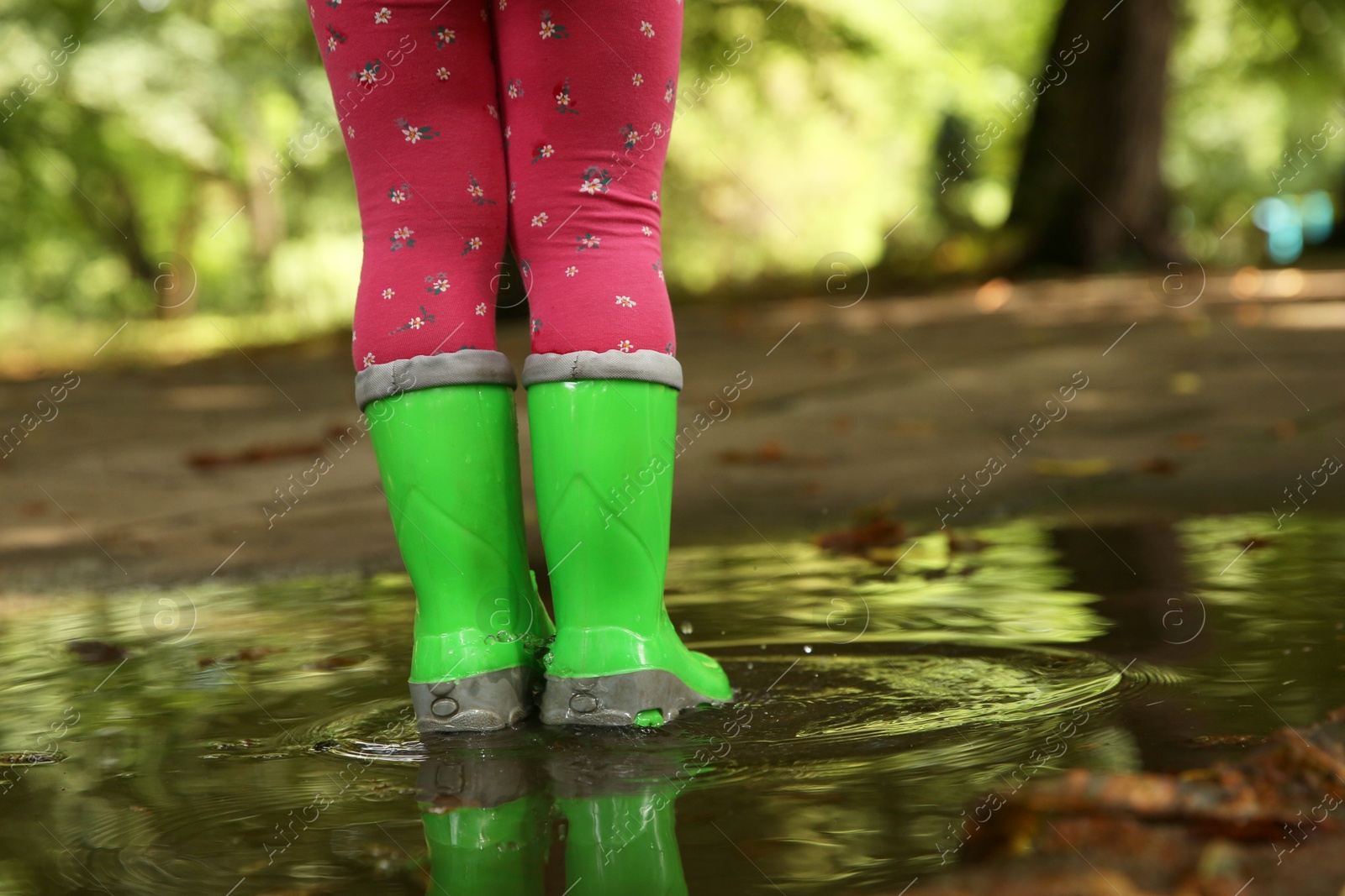 Photo of Little girl wearing green rubber boots standing in puddle outdoors, closeup