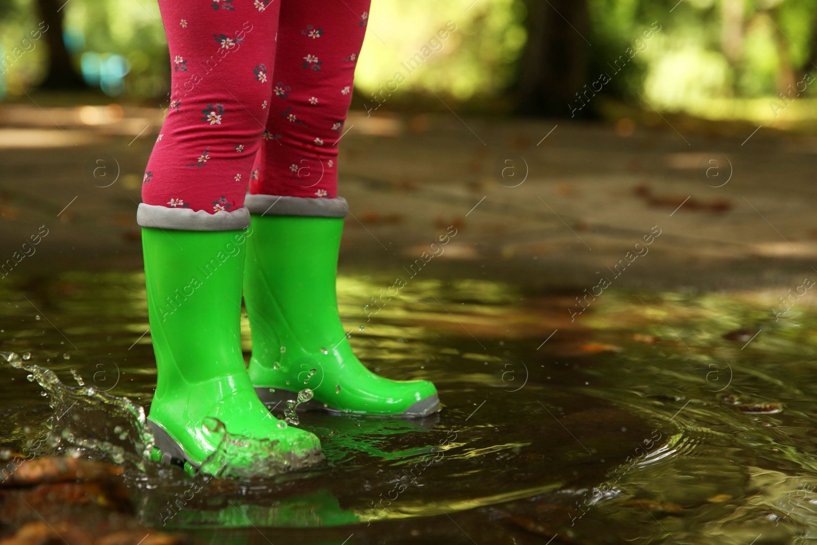 Photo of Little girl wearing green rubber boots standing in puddle outdoors, closeup. Space for text