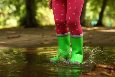 Photo of Little girl wearing green rubber boots standing in puddle outdoors, closeup. Space for text