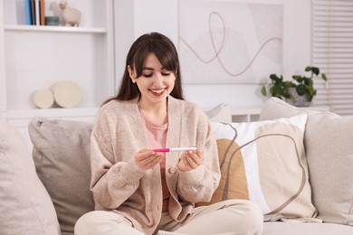 Happy young woman with pregnancy test on sofa at home