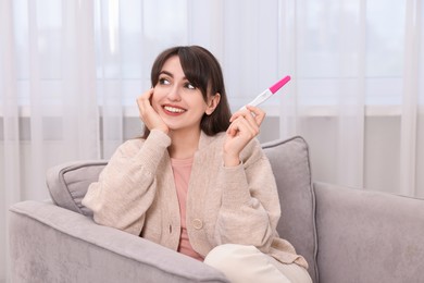 Photo of Happy young woman with pregnancy test on sofa at home