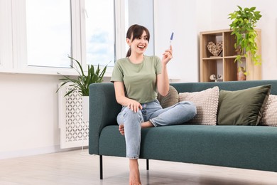 Photo of Happy young woman with pregnancy test on sofa at home