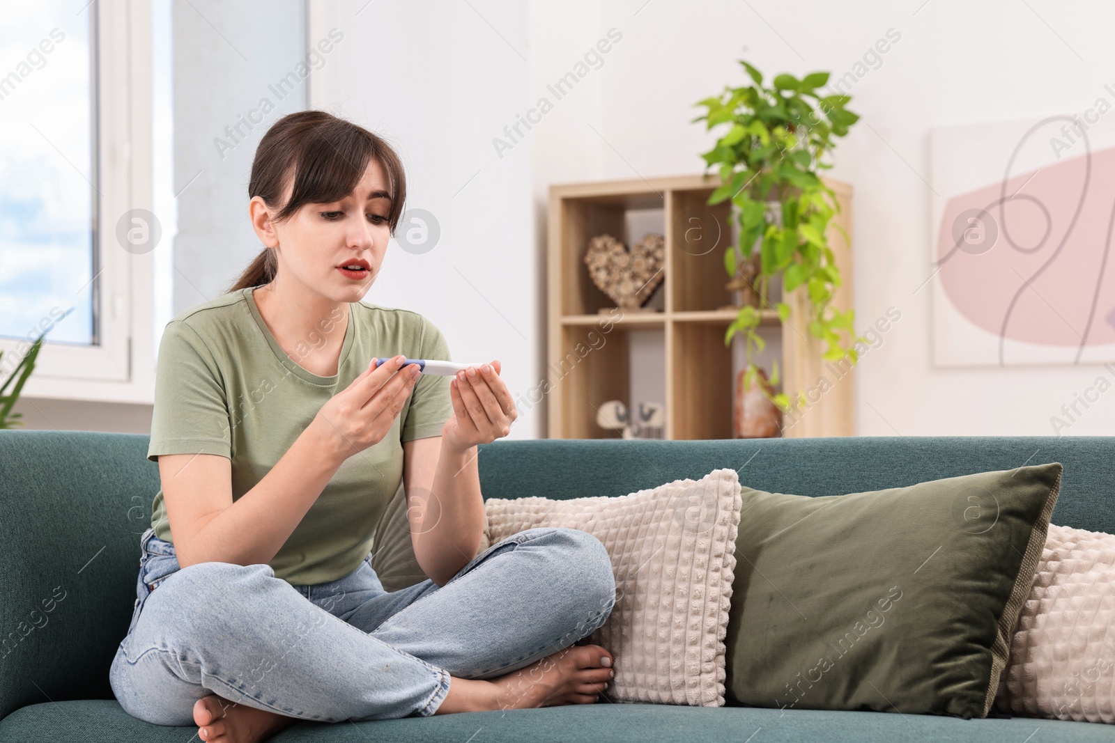 Photo of Worried young woman with pregnancy test on sofa at home