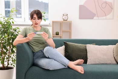 Photo of Worried young woman with pregnancy test on sofa at home