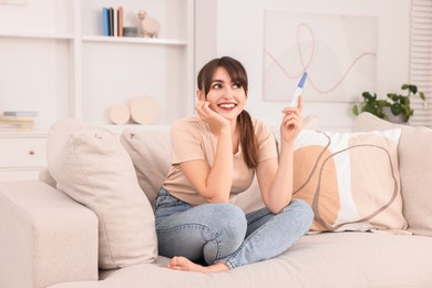 Photo of Happy young woman with pregnancy test on sofa at home