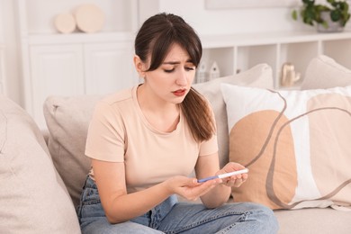 Photo of Worried young woman with pregnancy test on sofa at home