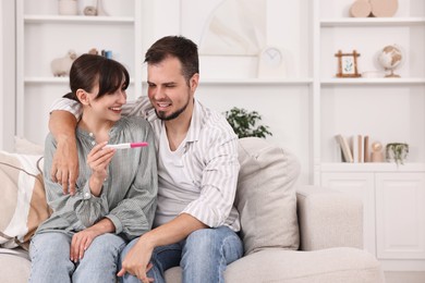 Happy young couple with pregnancy test on sofa at home, space for text