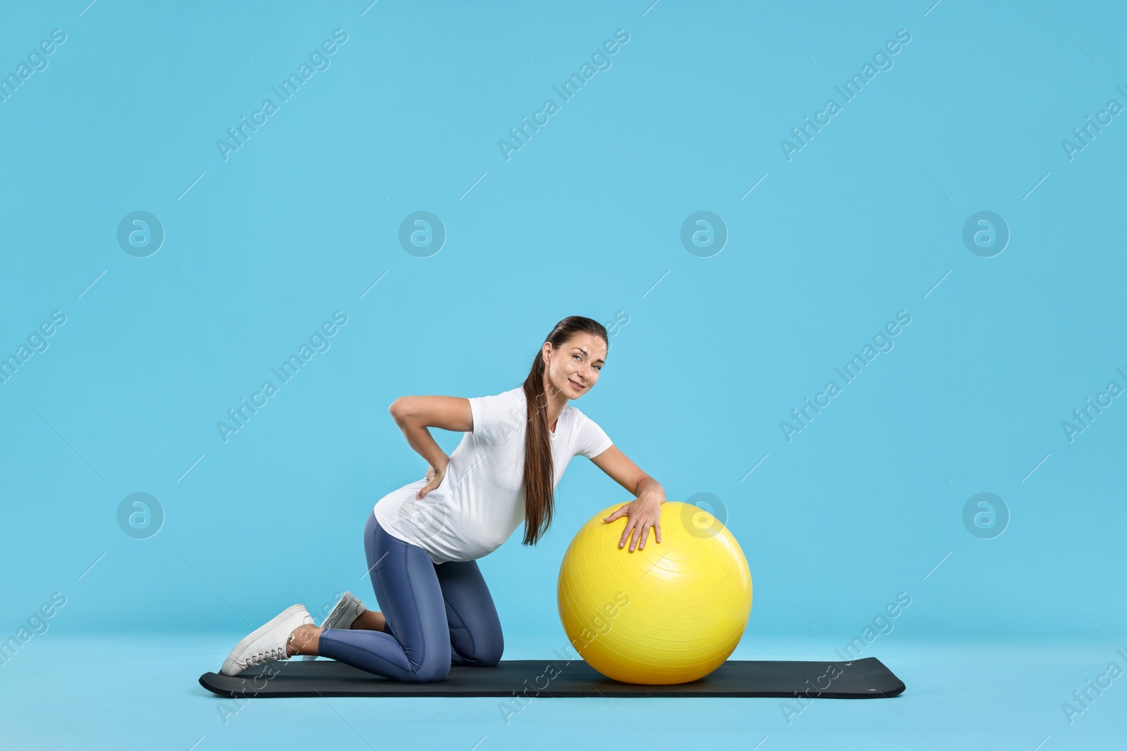 Photo of Beautiful pregnant woman doing exercises with fitball on mat against light blue background