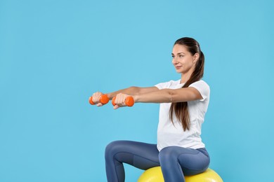 Beautiful pregnant woman with dumbbells doing exercises on fitball against light blue background