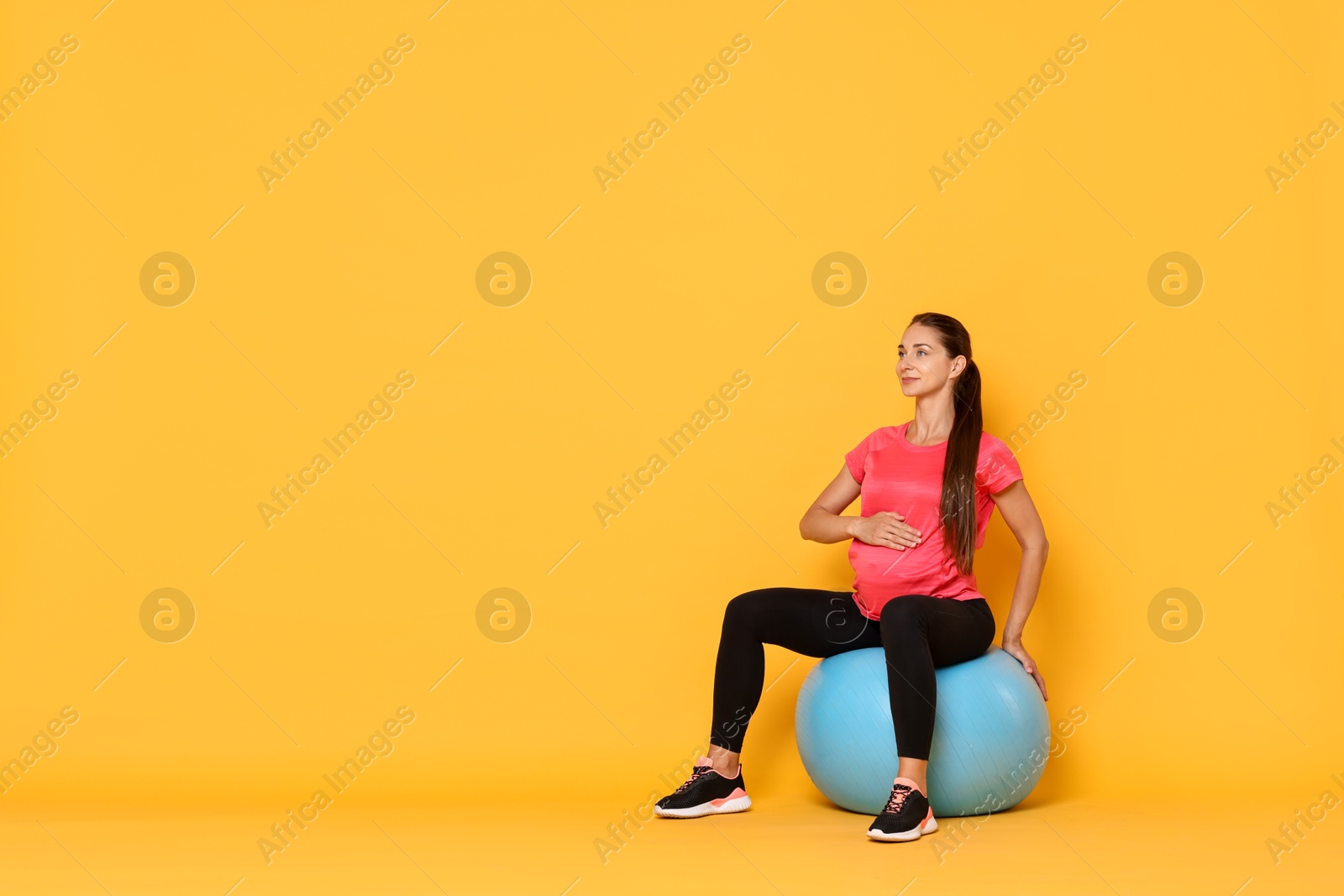 Photo of Beautiful pregnant woman doing exercises on fitball against yellow background, space for text