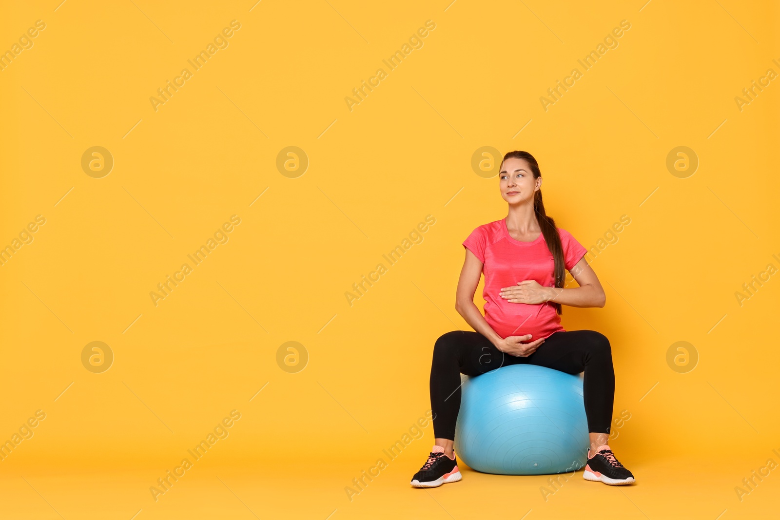 Photo of Beautiful pregnant woman doing exercises on fitball against yellow background, space for text