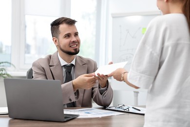 Photo of Smiling employee receiving envelope with salary from boss in office