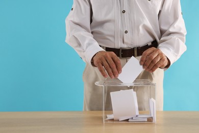 Referendum. Man putting his vote into ballot box at wooden table against light blue background, closeup