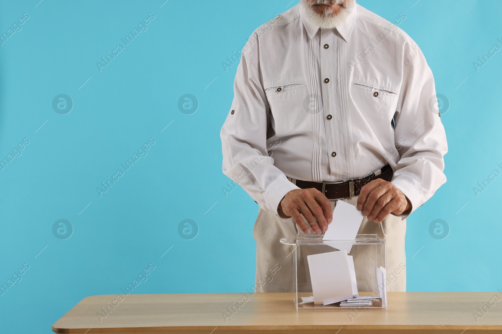 Photo of Referendum. Man putting his vote into ballot box at wooden table against light blue background, closeup