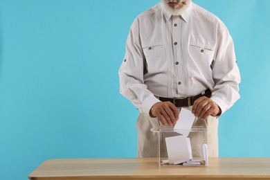 Referendum. Man putting his vote into ballot box at wooden table against light blue background, closeup