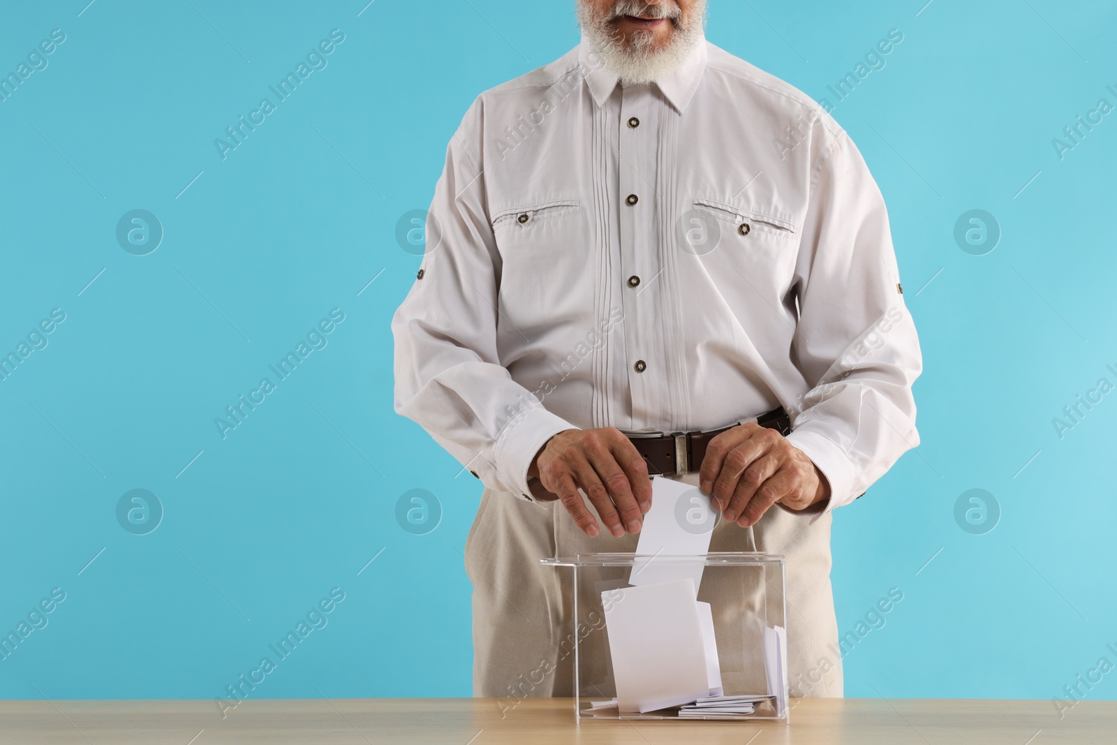 Photo of Referendum. Man putting his vote into ballot box at wooden table against light blue background, closeup