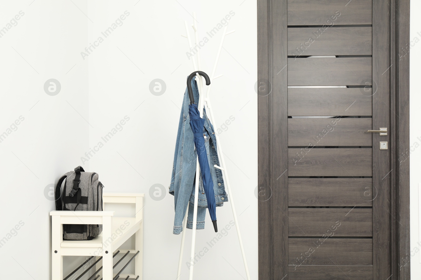 Photo of Blue umbrella and denim jacket on clothing rack near shoe storage bench with backpack in hallway