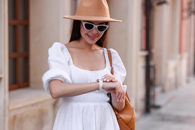 Smiling woman in stylish hat and sunglasses checking time on city street