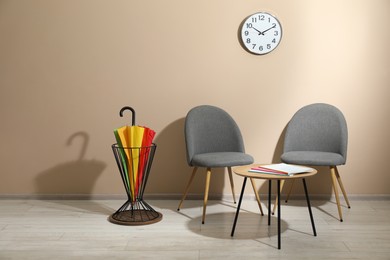 Photo of Bright umbrella in holder, coffee table and chairs near beige wall. Space for text