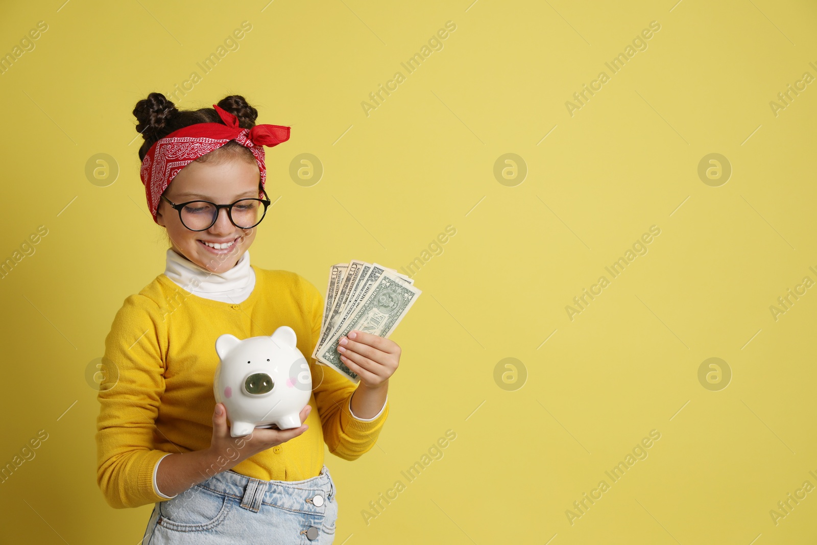 Photo of Pocket money. Cute girl with piggy bank and dollar banknotes on yellow background, space for text