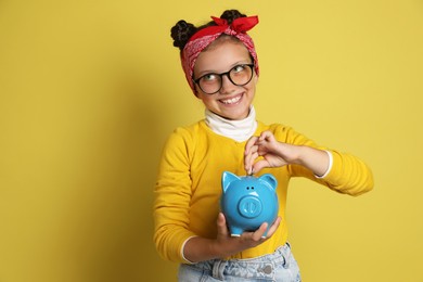 Photo of Pocket money. Cute girl putting coin into piggy bank on yellow background