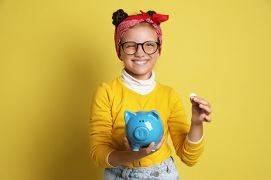 Pocket money. Cute girl putting coin into piggy bank on yellow background