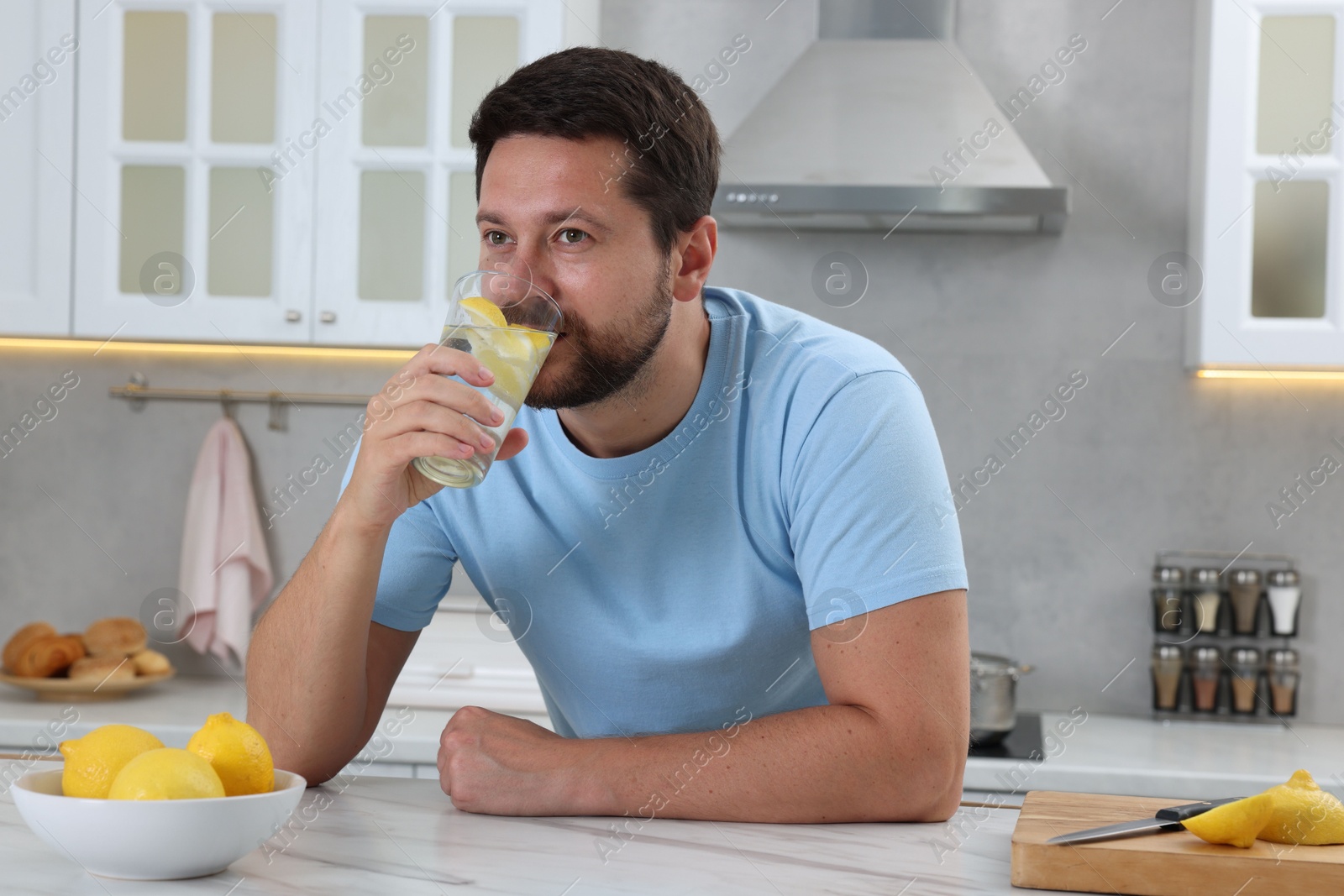 Photo of Handsome man drinking water with lemon at white marble table in kitchen