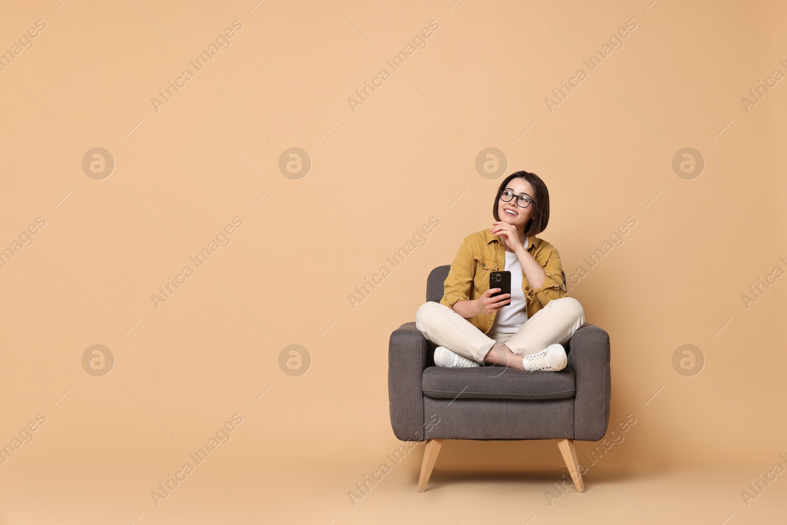 Photo of Smiling woman with smartphone sitting on armchair against beige background. Space for text