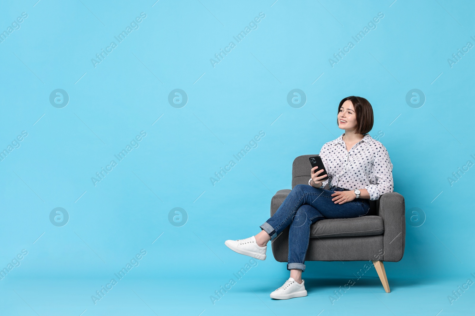 Photo of Smiling woman with smartphone sitting on armchair against light blue background. Space for text