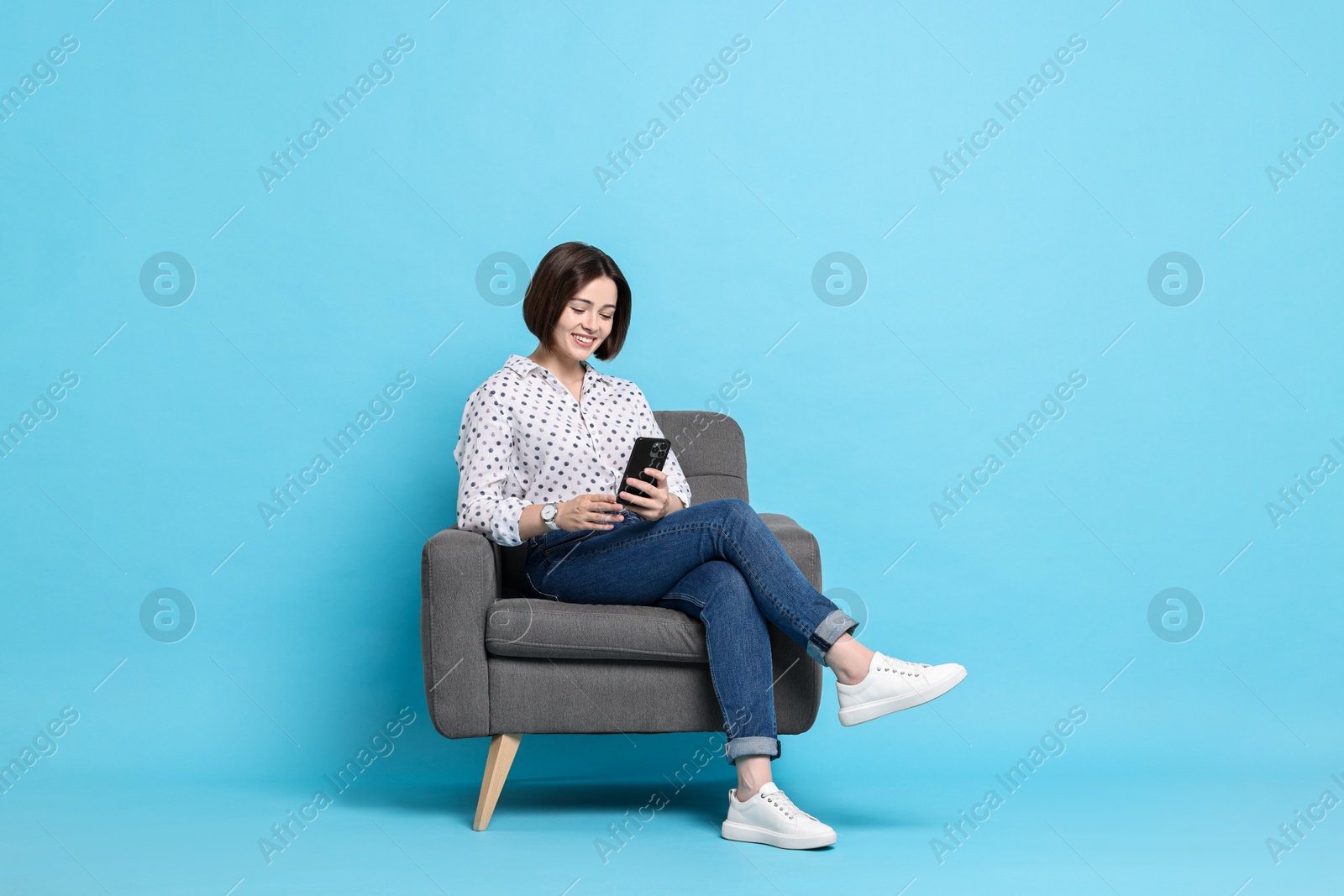 Photo of Smiling woman with smartphone sitting on armchair against light blue background