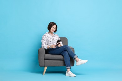 Photo of Smiling woman with smartphone sitting on armchair against light blue background