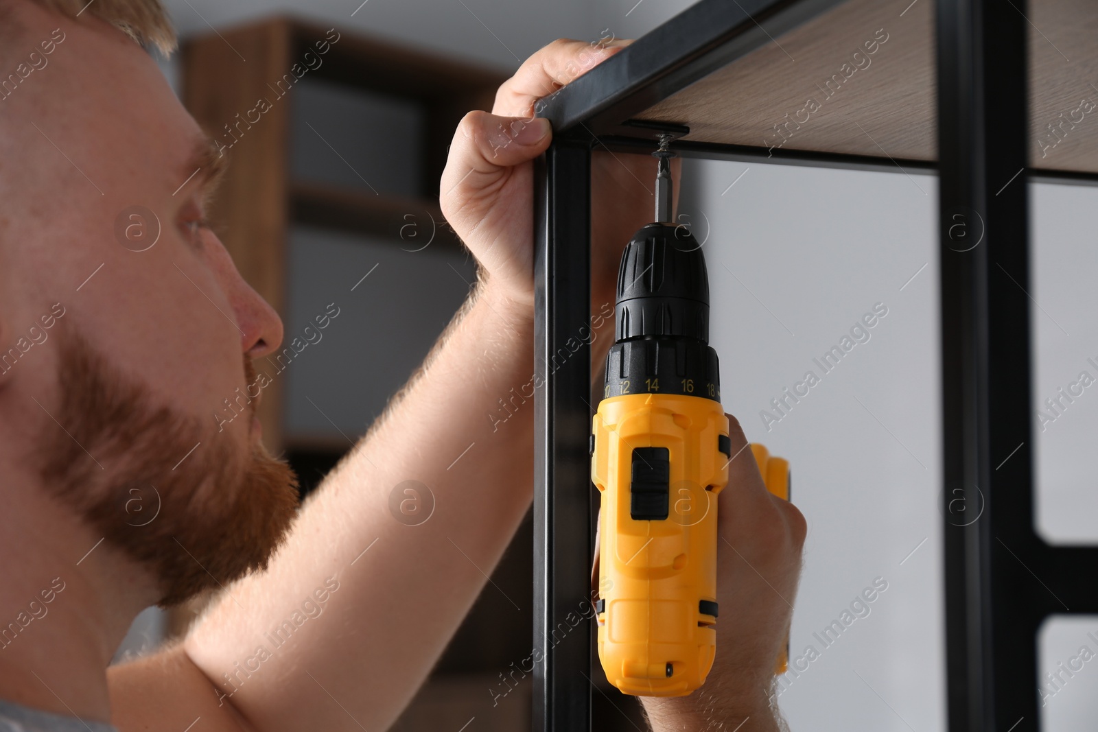 Photo of Man with electric screwdriver assembling furniture indoors, closeup