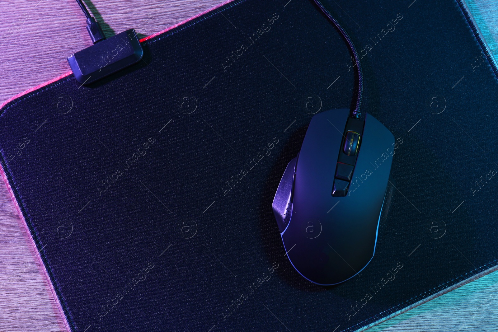 Photo of Black computer mouse and mousepad on wooden table in neon lights, top view